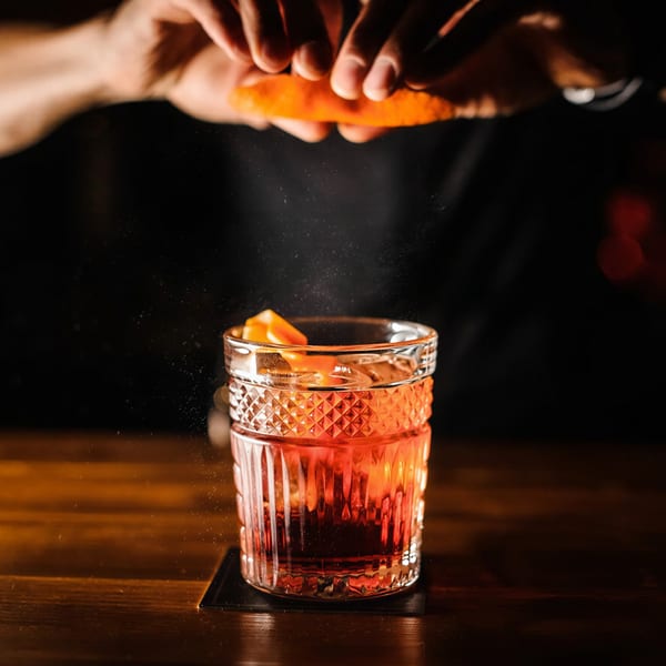 Bartender squeezing an orange peel over a vibrant red cocktail in a textured glass on a dark bar.