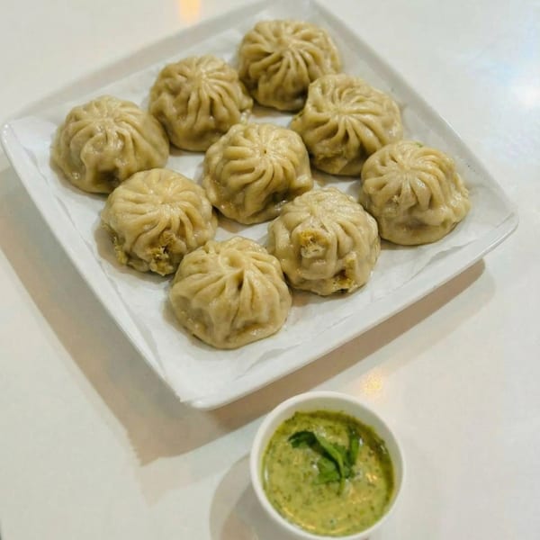 A white rectangular plate holds nine steamed dumplings next to a small cup of green dipping sauce.