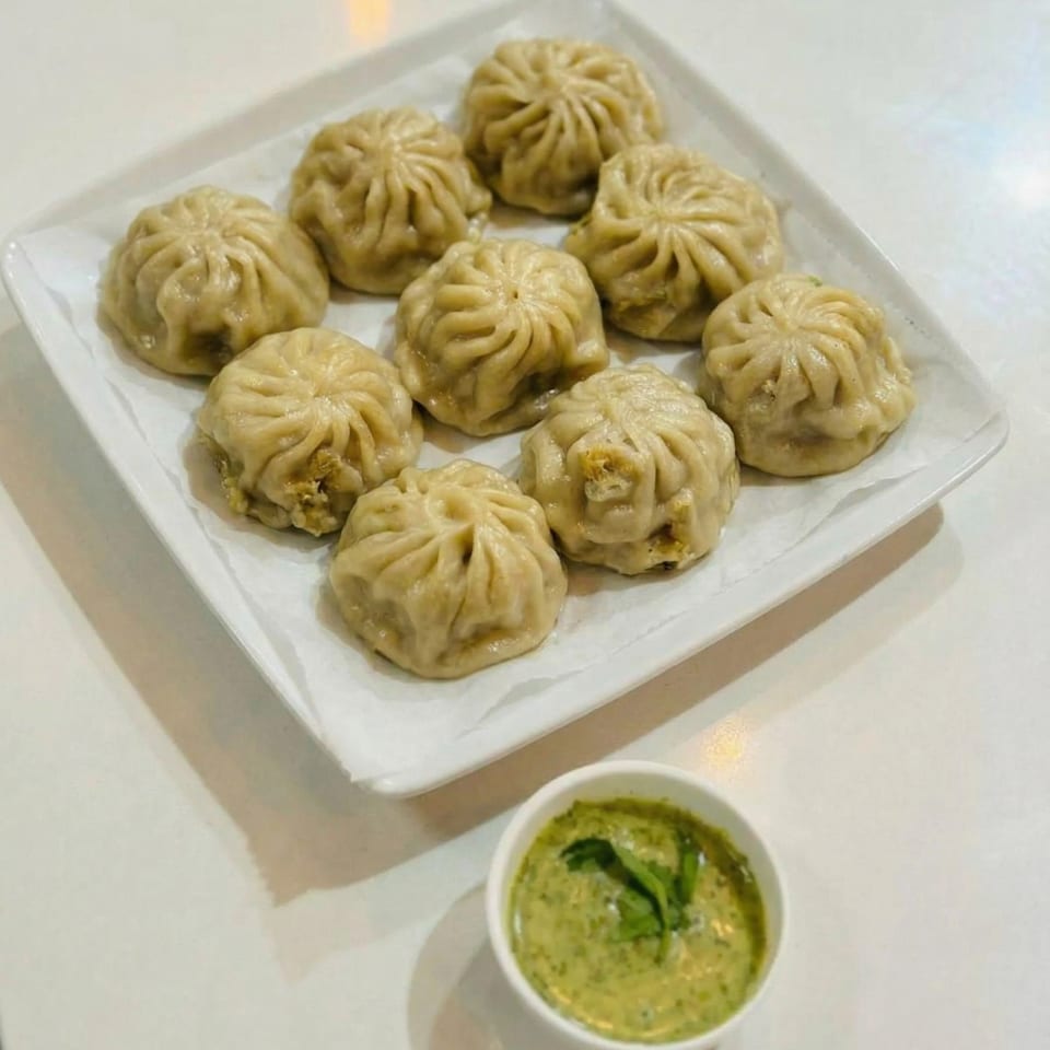 A white rectangular plate holds nine steamed dumplings next to a small cup of green dipping sauce.