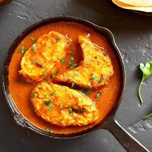Three pieces of fish in a rich orange curry sauce, garnished with fresh parsley in a black pan.