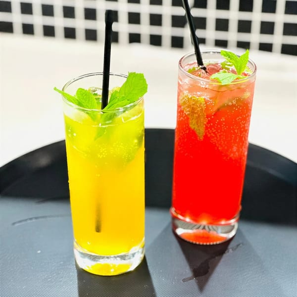Two tall glasses of colorful, bubbly drinks with mint and straws on a dark tray.
