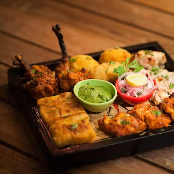 A wooden tray filled with various Indian appetizers, including chicken lollipops, paneer, and sauces.
